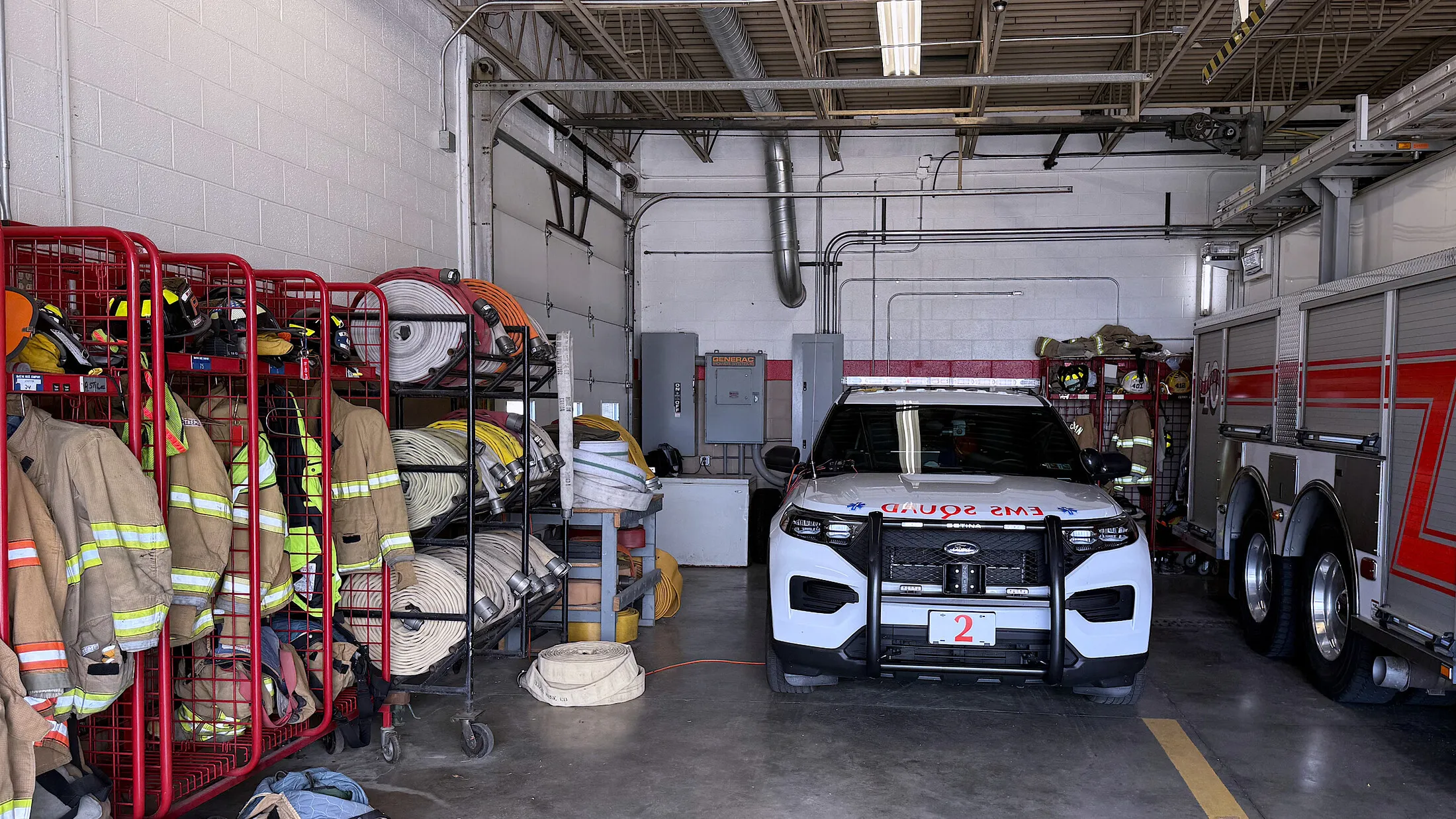 Firefighting coats, helmets and rolled fire hose on steel racks along the wall of the McKean Hose Company truck bay, with a department vehicle and a fire engine parked in the bays alongside