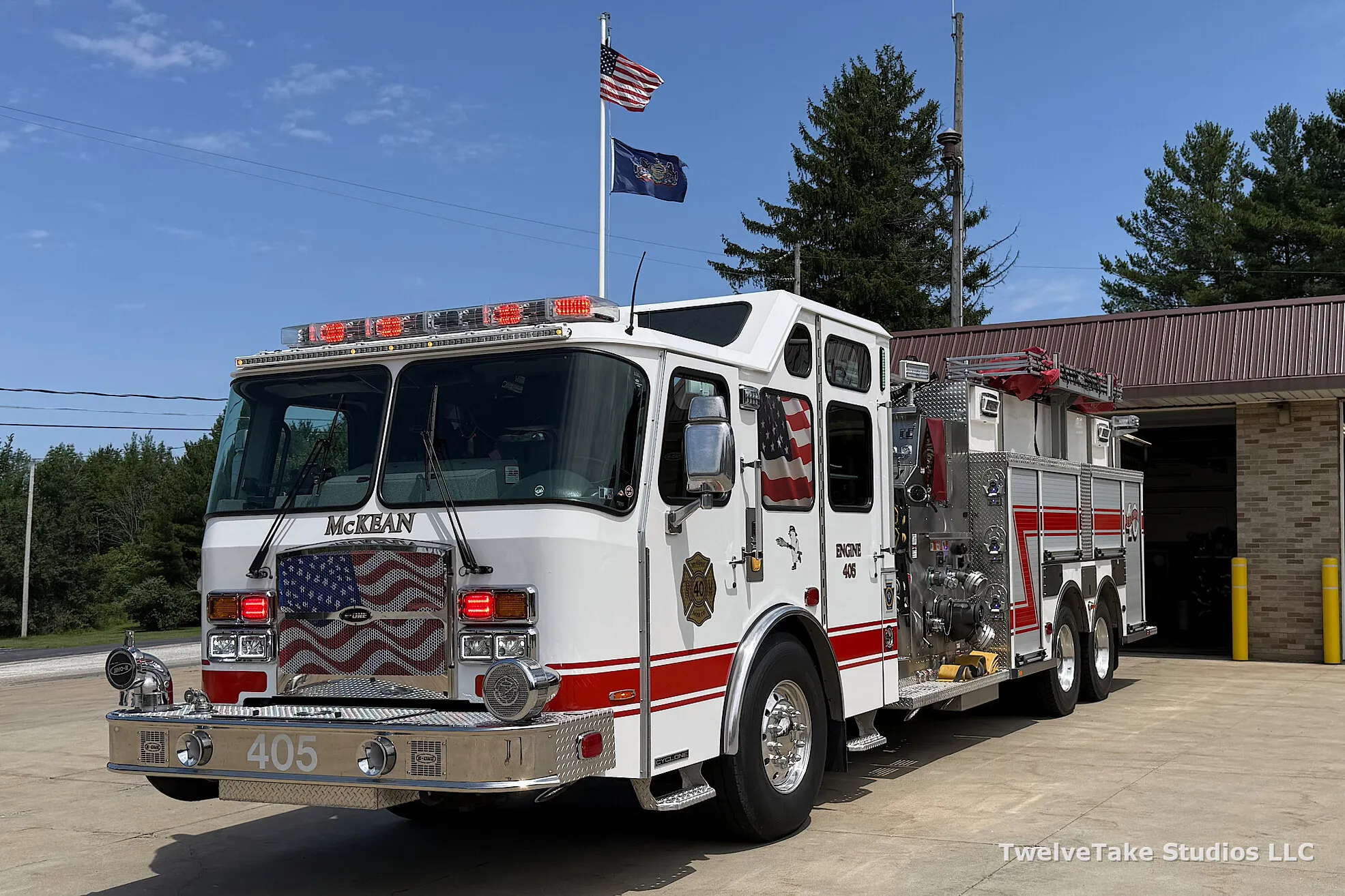 Engine 405 parked on the pavement outside the McKean Hose Company station with its red warning lights lit and the American and Pennsylvania flags flying behind it.