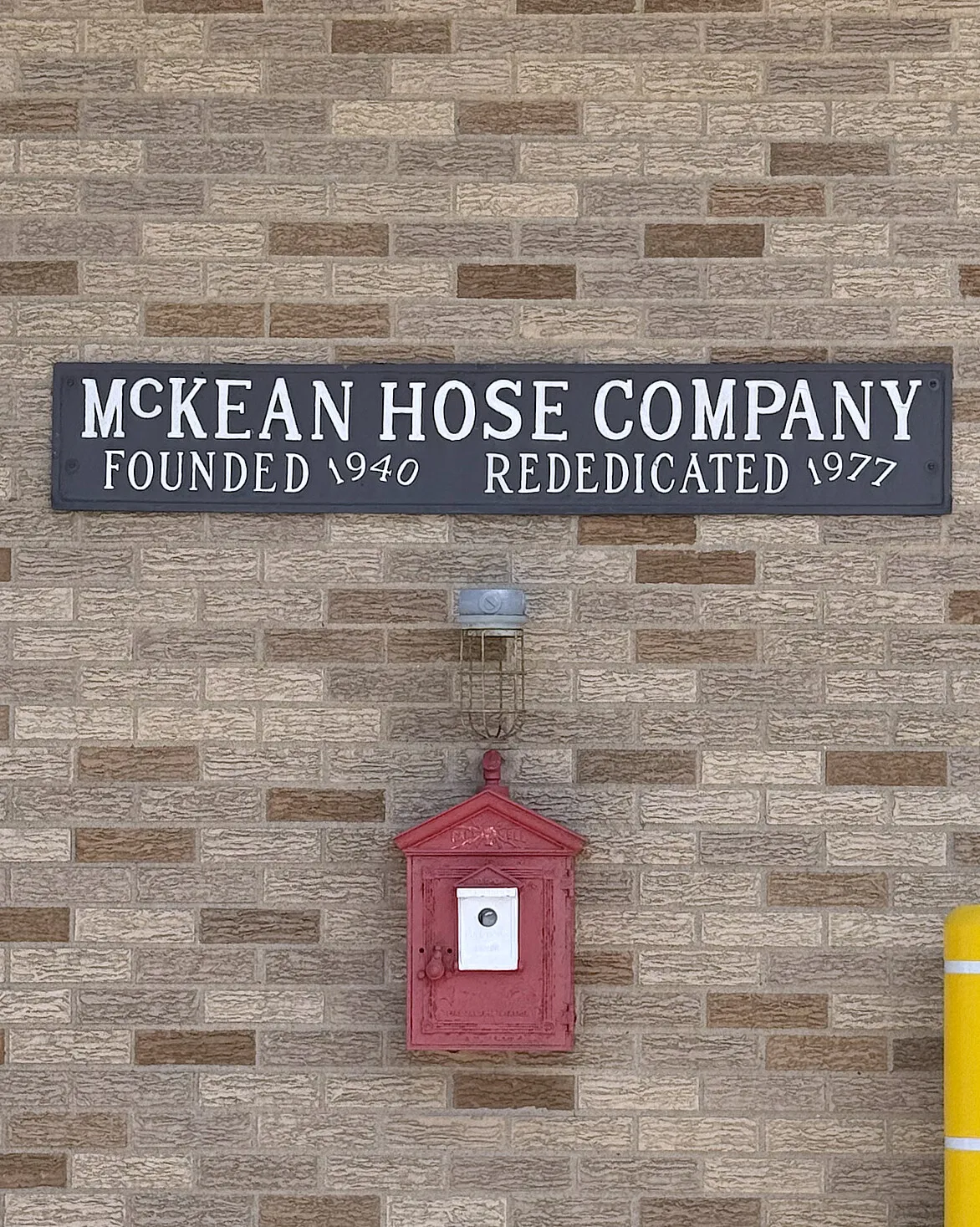 A cast metal plaque on the station's brick wall reading McKEAN HOSE COMPANY, FOUNDED 1940, REDEDICATED 1977, mounted above an old red cast-iron fire alarm call box.
