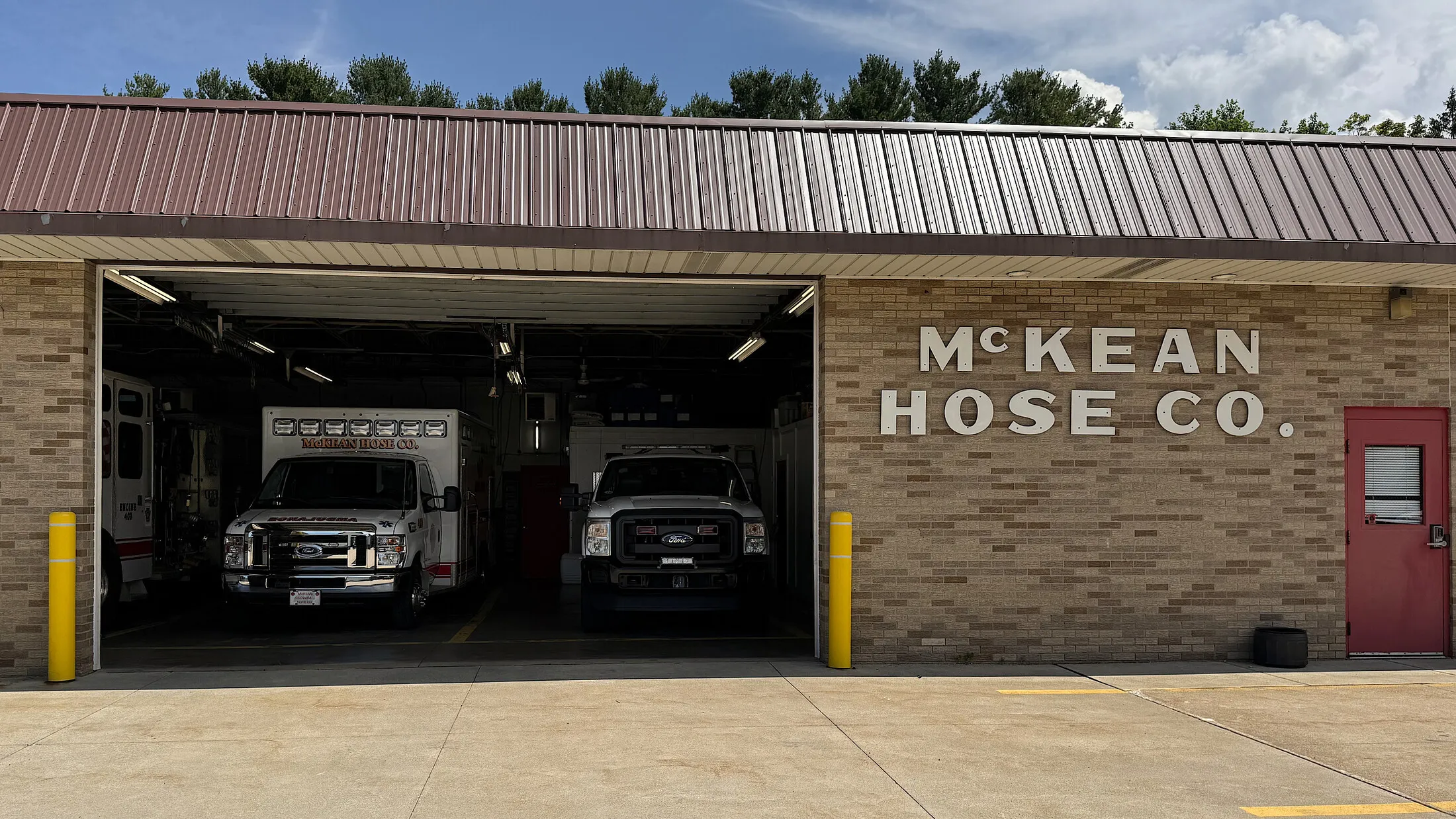 The McKean Hose Company station: a brick building with a dark metal roof, bay doors raised with an ambulance and a pickup inside, McKEAN HOSE CO. spelled out in white letters on the brick front, and a red side door at the right.