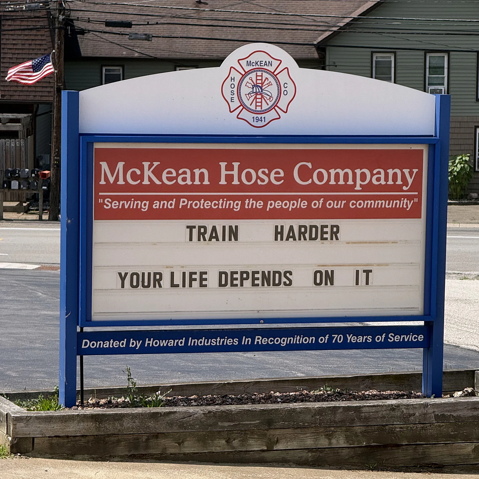 The roadside sign outside the station, blue framed, topped with the department crest and reading McKean Hose Company above a changeable letterboard that says TRAIN HARDER, YOUR LIFE DEPENDS ON IT.